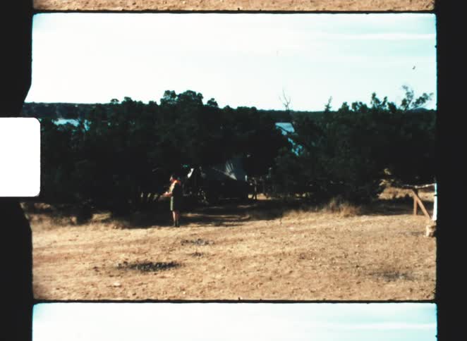 A wide shot of a dusty campsite shows several large tents among the woods near a body of water with people walking around the foreground
