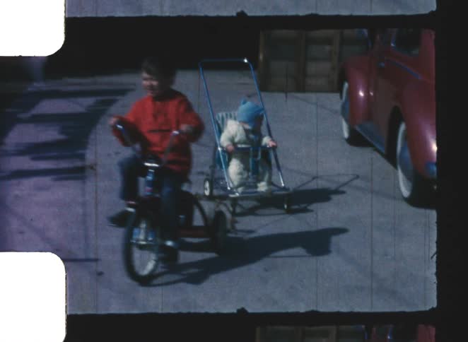 A young boy riding his bike in circles on the driveway at midday pulling a baby in a stroller attached behind him with a red car parked off to the side