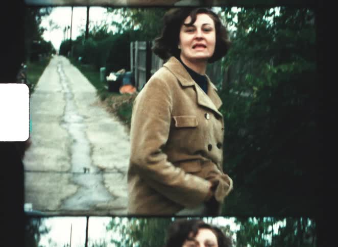 A woman wearing a tan jacket and white pants gets up from a cart then turns to smile and adjust her hair while looking directly at the camera with two other people in the background
