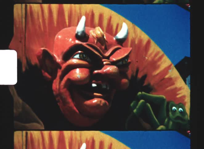 A tight closeup shot of a large brightly painted red devils head sculpture featuring menacing eyes and white horns is shown with a small green spider figure visible near its right shoulder