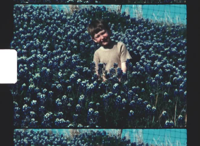 A young boy with short brown hair is seen smiling directly at the camera while sitting amid a dense patch of bluebonnets on a hillside with a barbed wire fence visible behind him