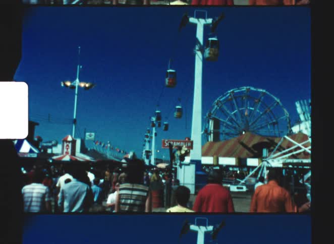A crowd of people gathers at an outdoor fair with a ferris wheel and a sky ride with colored gondolas under a deep blue sky captured in a vintage 8mm film style