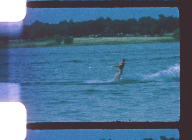 A person in an orange life vest is seen water skiing across a large choppy blue lake being pulled by a speeding boat that exits the frame leaving a wide white wake while a forested shoreline and a distant sailboat are visible in the background