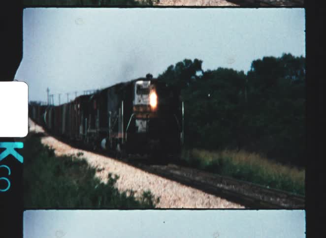 Captured on grainy vintage stock a diesel locomotive powers down a rural track with its bright headlight beaming steadily approaching the camera through a nostalgic sunwashed landscape