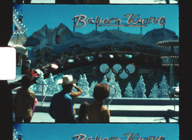 A group of people watching the bayern kurve amusement park ride which features a fastmoving alpinethemed backdrop with olympic rings decor under a bright blue sky