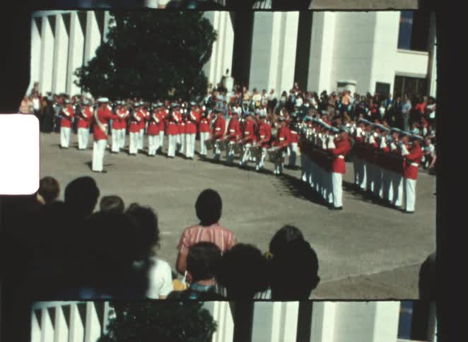 Military band in red uniforms performs in formation outdoors in front of a gathered audience near an institutional building