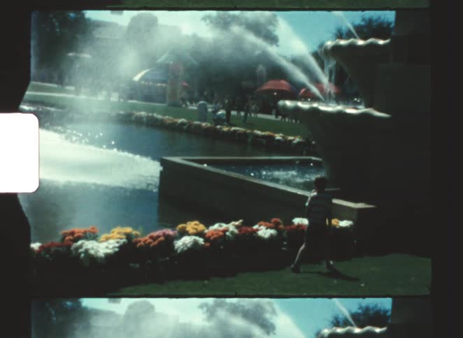 A child walking across a grassland toward a water fountain with flowers lining the sides at midday