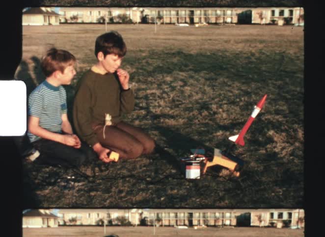 Children prepare model rocket launch on grassy field near residential houses