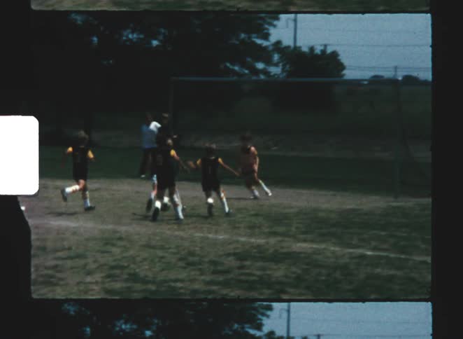 A group of children in uniforms plays a fast moving soccer game on a grassy outdoor sports field in vintage film footage