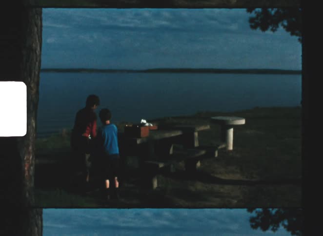 A mother and child setting up a picnic on an outdoor bench by the riverbank at midday