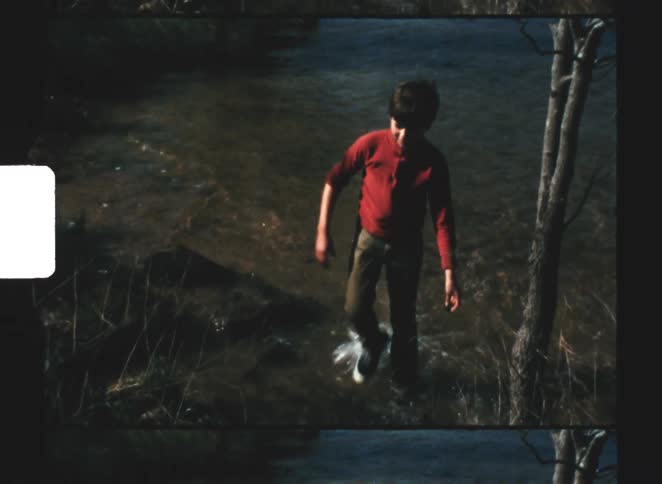 A boy in a red shirt and pants slowly wades toward the camera through the shallow flowing water of a stream surrounded by dark foliage and tree trunks