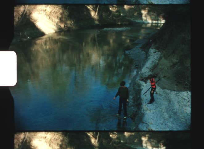 Two young kids having fun together on a riverbank picking up sticks at midday