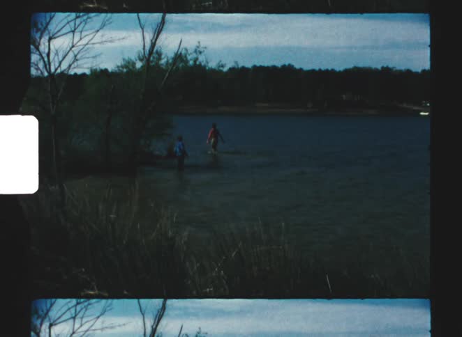 A small body of water bordered by tall grass and trees shows two distant people standing near the shoreline under a darkened blue sky