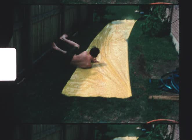A teenage boy sliding down a wet backyard slide at midday while another kid watches both shirtless