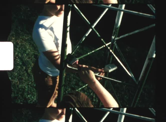 Two children are seen from above in a vintage style home video with film grain as one child paints a white metal structure outdoors