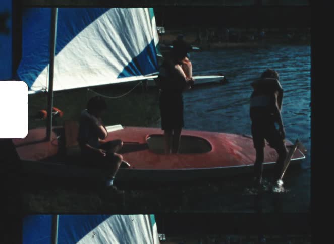 Three boys are standing near a small sailboat with a blue and white sail near a body of water in vintage film