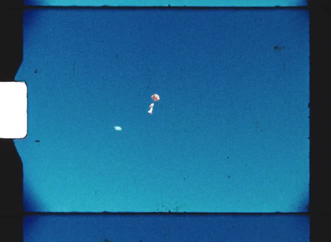 A parachute with a small toy figure descends slowly against a vast clear blue sky in vintage film footage
