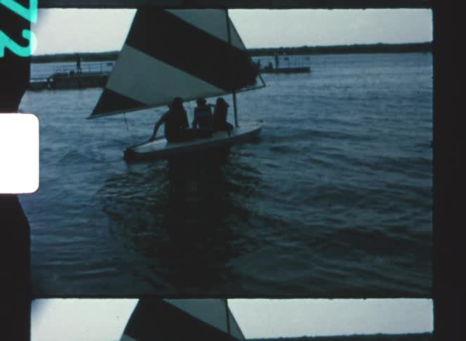 Three people in a small sailboat sailing on a lake near a pier