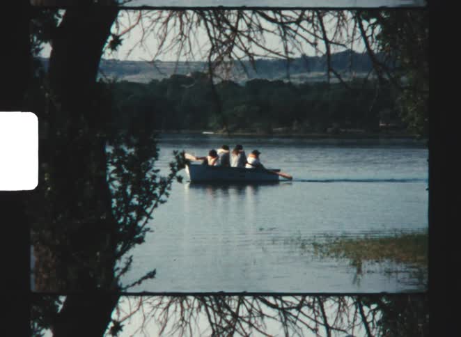 Framed by dark foreground trees four people are visible paddling a small rowboat across a wide calm body of water toward a distant shore of dark wooded hills