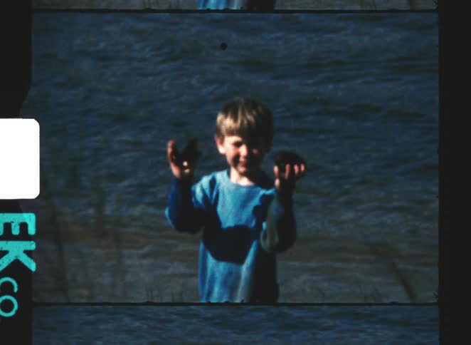 A smiling young boy in a blue sweater stands waistdeep in rippling water proudly holding up two large dark rocks for the camera