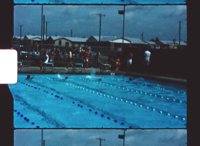 People watch swimmers compete in an outdoor pool on a cloudy day