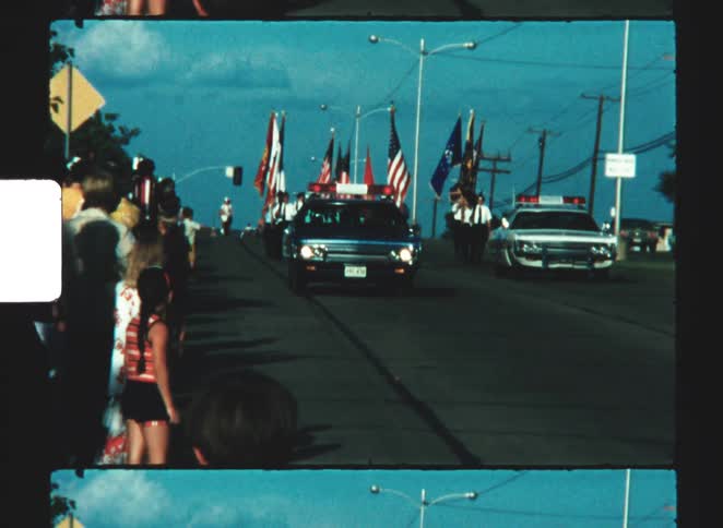 A parade moves down the street led by police cars with spectators along the sidewalks