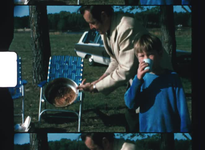 An older man in a tan jacket is cooking food in a pan over an outdoor fire then proudly shows the prepared meal to a young boy who sips from a white cup