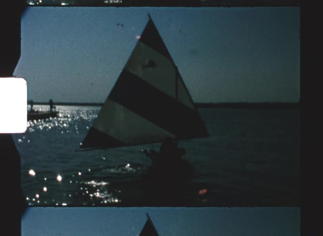 A small sailboat carrying two people is silhouetted against the dark water sailing towards the setting sun whose intense light glints brightly off the surface