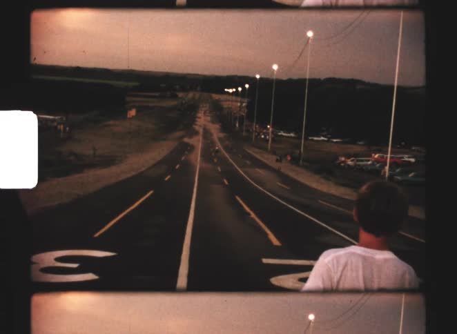 A kid standing in the middle of the road watching another kid ride a bike down a hilly road with a streetlight on the right side at sundown