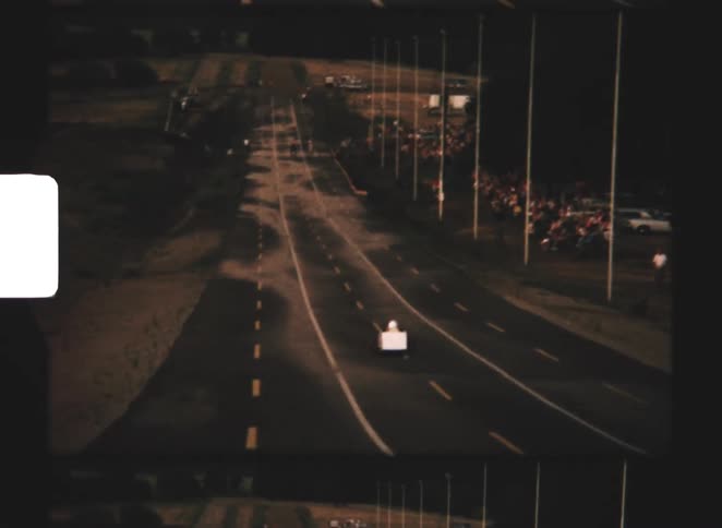 White soapbox car with red wheels rolls down paved track in front of vintage car lot