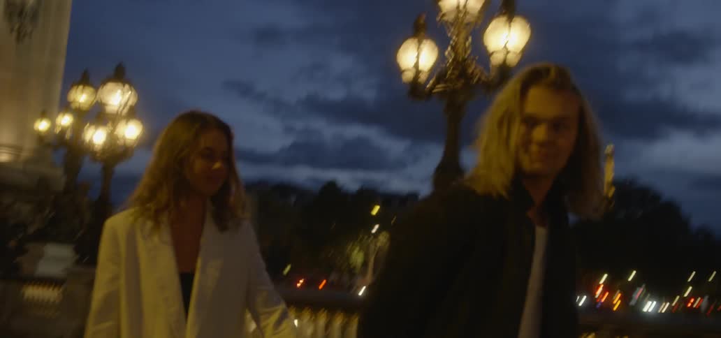 A man and woman holding hands walk along a well lit bridge at dusk