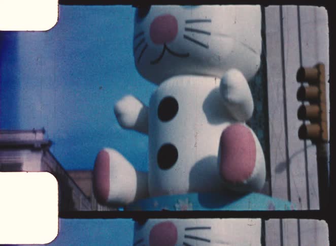 A giant inflatable white easter bunny with pink ears and nose is lowered down past a traffic light in a vintage city parade