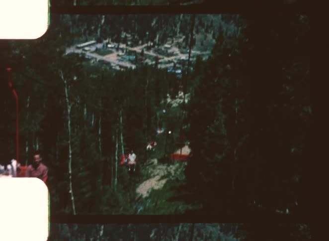 A downhill movement from a chairlift passing over a dense pine forest and capturing people seated on the lift chairs with a small town visible in the distance below