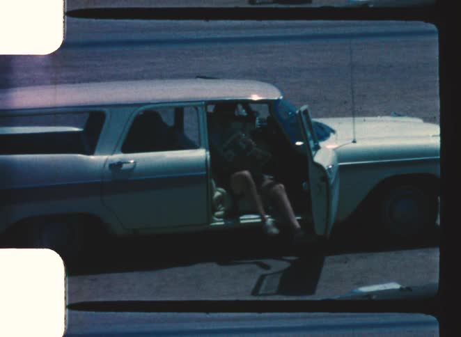 A white and teal vintage station wagon is parked as a person in shorts steps out from the drivers side door onto a paved road