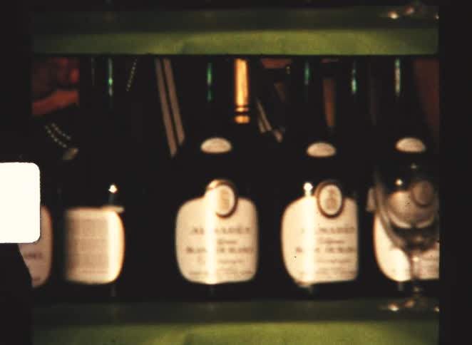 Jugs of alcohol and a wine barrel sit on the shore near the water while wine bottles stand on a shelf