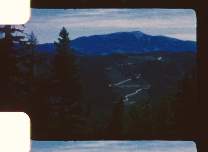 A wide shot of a valley road winding up a forested mountain range under a blue overcast sky