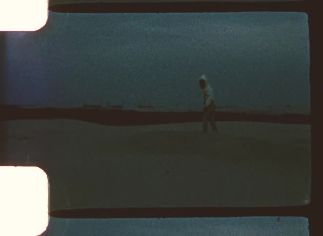 A person in a hooded white garment walks slowly across a large flat and dark expanse under a blue gray sky