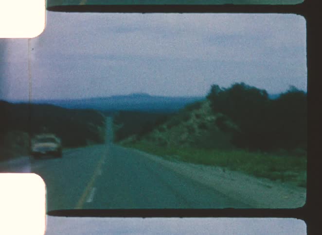 Vehicle travels twolane road between greenery and mountains under cloudy sky