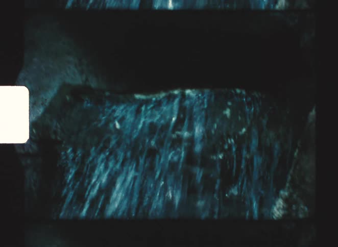 A low angle shot with vintage film grain shows water cascading over a large rock formation