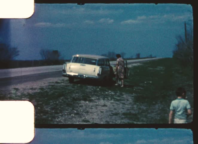 A lightcolored station wagon is parked on a gravel roadside under a dark blue sky with two people walking near the open rear door and another walking away along the edge of the road amid sparse vegetation