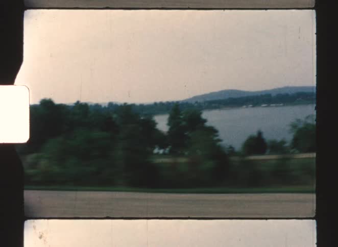 A view from a moving vehicle shows a blurred foreground of road and foliage with a lake and distant hills visible
