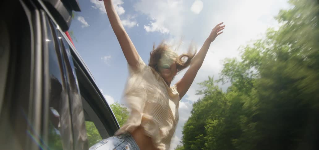 A woman leans out the window of a moving vehicle and lifts her arms