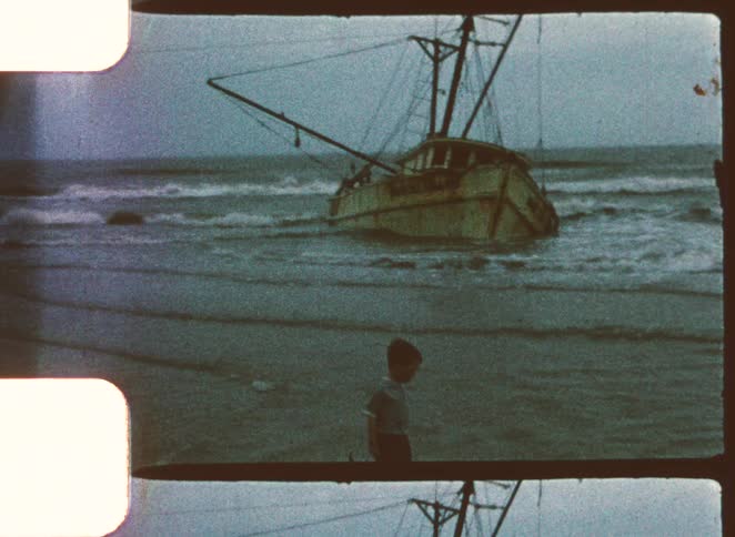A kid walking along the beach into the shallow water with a wrecked boat sitting offshore in the background as waves crash against it at midday