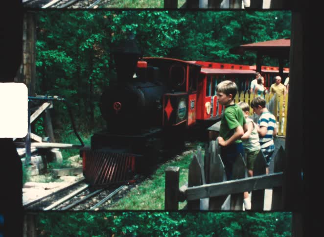 Small train with black engine and red car arrives at station in wooded area as children watch