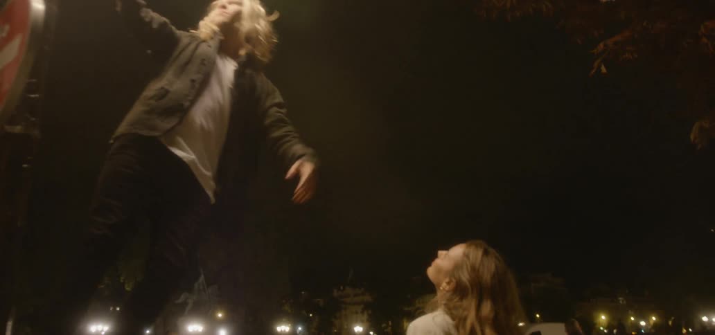 A man hangs from a lamp post and a woman stands on the sidewalk in a city at night