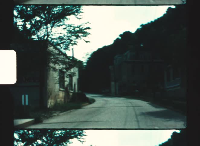 Vehicle drives through a town bordering a forested hill during the daytime