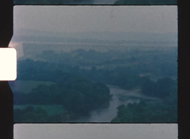 A massive white dam spanning a valley behind a darkforested foreground the camera pans down to reveal a winding river and misty rolling hills in the distance