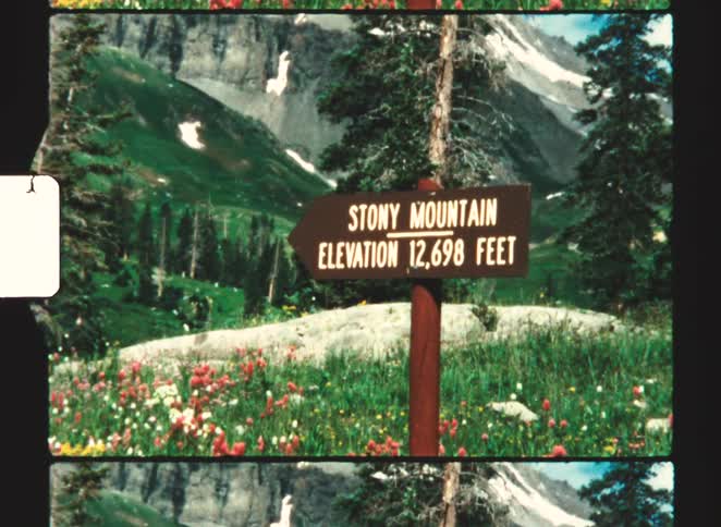 A red wooden road sign pointing left surrounded by grass with the text stony mountain elevation 12698 ft and a mountain range visible in the background at midday