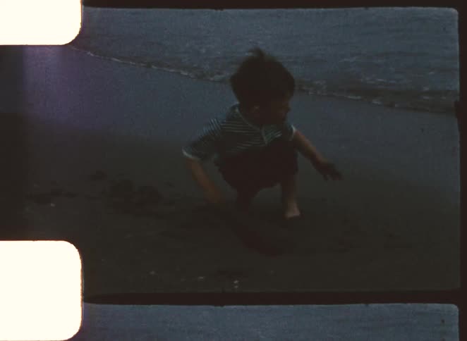 Child in striped shirt plays in beach sand near shoreline at dusk