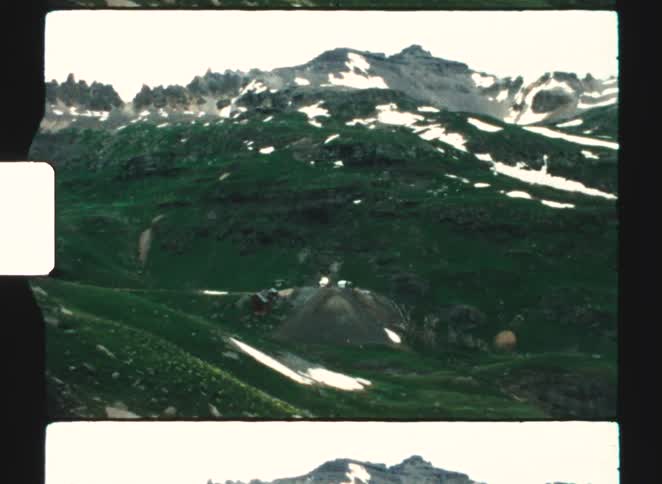 A majestic mountain range with steep rocky peaks and slopes covered in patches of green grass and remnants of snow featuring a small structure and road in the valley below